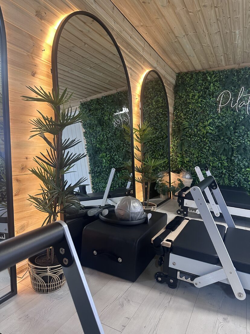 A reformer machine beside a softly lit arched mirror and the green Pilates wall